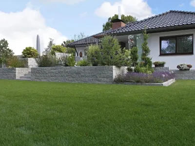 Eine massive Stützmauer aus Granitmauersteinen fängt ein erhöhtes Beet vor einem grünen Rollrasen ab.