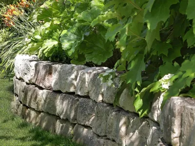 Eine solide Trockenmauer aus Naturstein stützt ein üppig bepflanztes Beet entlang der Rasenfläche.